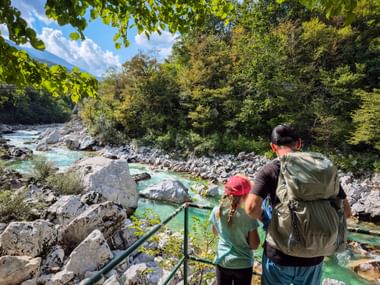 Zwei Wanderer mit Rucksäcken blicken vom Aussichtspunkt Trnovo auf die türkisfarbene Soča in den Julischen Alpen, umgeben von Flussbett und Wald.