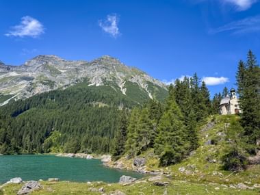Kleine Kapelle auf einem bewaldeten Hügel neben dem türkisfarbenen Obernbergersee, mit felsigen Berggipfeln und Nadelwald unter blauem Himmel.