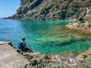 Hiker with blue backpack sitting on rocky shore in Bonassola, overlooking turquoise Mediterranean bay with steep cliffs in Cinque Terre.