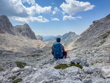 Wanderer mit blauem Rucksack sitzt auf felsigem Gelände in Vršaki, Julische Alpen, mit Blick auf Bergtal mit dramatischen Gipfeln unter blauem Himmel.