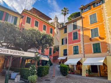 Colorful Mediterranean buildings in yellow, orange, and pink surround a cobblestone square in Portofino with luxury shops and a church tower.
