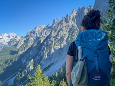 Hiker with blue Vaude backpack viewing the jagged Gosaukamm mountain range in Salzkammergut under clear blue sky.