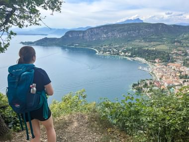 Wanderer mit blauem Rucksack blickt vom erhöhten Wanderweg auf den Gardasee und den Ort Garda mit Bergen und blauem Wasser.