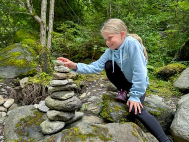 Young girl in blue hoodie crouching on moss-covered rocks, placing stones on a cairn in a lush green forest setting in Passeier Gorge.