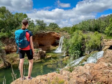 Female hiker with blue backpack standing on rocky cliff overlooking Pomarinho waterfall in Algarve, surrounded by green vegetation under cloudy sky.