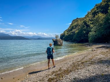 Wanderer mit Rucksack am Kieselstrand von Lido di Manerba, Gardasee. Türkisfarbenes Wasser, Felsformation und bewaldeter Hang sichtbar.
