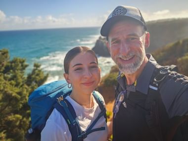 Vater und Tochter mit Rucksäcken auf Küstenpfad bei Almograve, Alentejo. Blaues Meer und Klippen hinter ihnen unter sonnigem Himmel.
