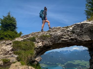 Female hiker with backpack standing on natural rock arch overlooking alpine lake and mountains in Salzkammergut region.