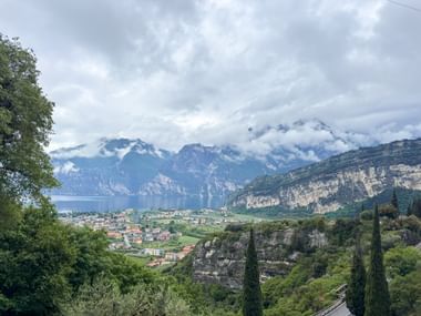 Panoramablick von Nago über Torbole und den Gardasee mit teilweise wolkenverhangenen Bergen, Zypressen und Vegetation im Vordergrund.