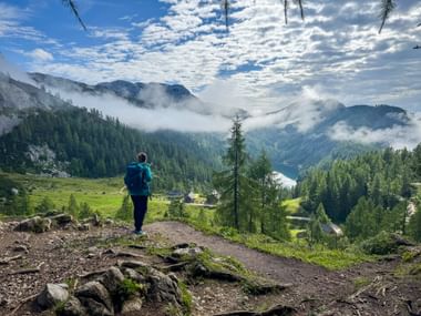 Wanderin mit blauem Rucksack auf Bergweg der Tauplitzalm mit Blick auf den Steirersee, umgeben von bewaldeten Bergen und Morgennebel.