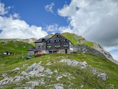 Leutkircher Hütte, a traditional alpine hut with blue shutters on a grassy hillside, surrounded by rocky peaks under a partly cloudy sky.