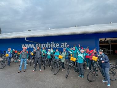 Large group of cyclists with bikes and yellow bags posing in front of blue Eurobike building during company trip along the Danube.