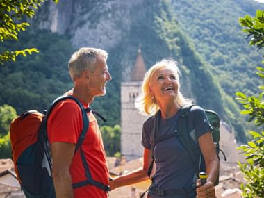 Two smiling hikers with backpacks in Gemona, Italy. Behind them, a church tower and castle ruins on a green mountainside are visible.