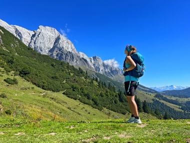 Wanderin mit Rucksack steht auf grüner Almwiese und blickt auf dramatische Kalksteingipfel in den Bayerischen und Salzburger Alpen unter blauem Himmel.
