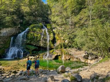 Zwei Wanderer mit Rucksäcken und Wanderstöcken stehen an einem klaren grünen Becken am Virje-Wasserfall, umgeben von moosbewachsenen Felsen.