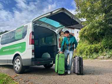 Man in turquoise jacket standing with green and black suitcases at open rear door of white-green Eurohike luggage transfer van.