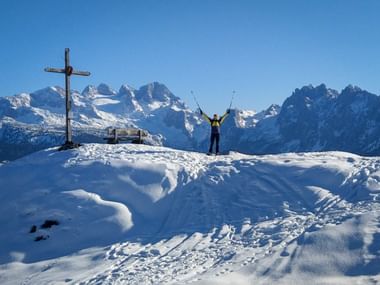 Wanderer mit erhobenen Skistöcken am verschneiten Bodenberg-Gipfel mit Holzkreuz, mit Blick auf schneebedeckte Berggipfel unter blauem Himmel.