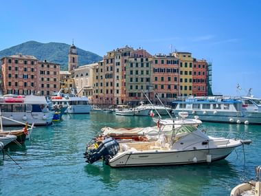 Harbor of Camogli with boats moored in turquoise water, colorful buildings along the waterfront, church tower, and mountain in background.