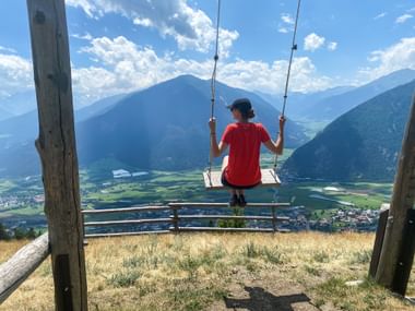 Person in red shirt swinging on wooden swing overlooking Vinschgau valley with mountains, green fields, and village of Schluderns below.