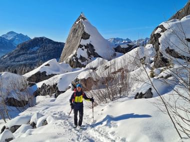 Winterwanderer mit Rucksack und Stöcken auf verschneitem Weg nahe markanter Felsformation Predigtstuhl mit Bergpanorama unter blauem Himmel.