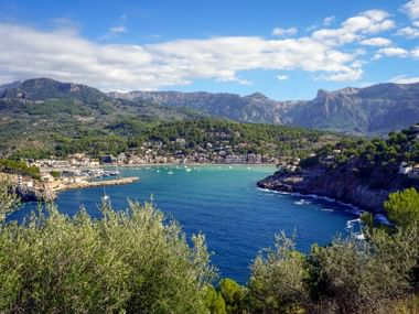 Panoramic view of Port de Sóller bay with turquoise water, harbor, coastal town, and Tramuntana mountains under blue sky with white clouds.