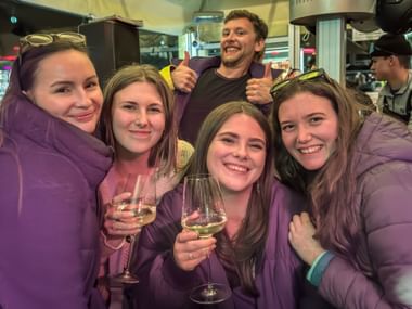 Gruppe von fünf Personen in lila Winterjacken lächelnd beim Wintersporttag 2026 Après-Ski, mit Weingläsern in Innenräumen.