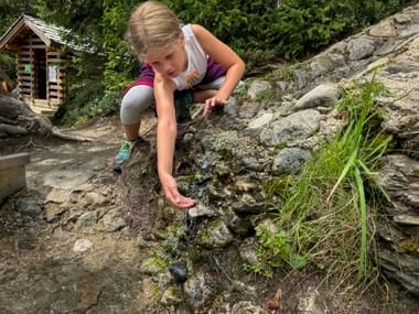 Young girl crouching on rocks beside a mountain stream, reaching toward the water. A wooden shelter is visible in the background among trees.