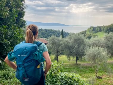 Wanderin mit blauem Rucksack blickt vom Hang mit Olivenbäumen, Zypressen auf den Gardasee mit Bergen in der Ferne unter bewölktem Himmel.