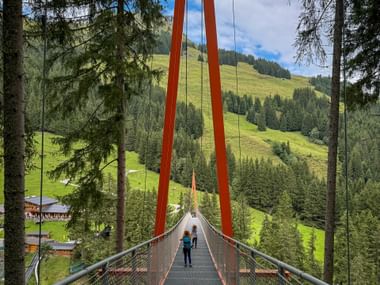 Hikers crossing a suspension bridge with orange towers on the Baumzipfelweg in Saalbach-Hinterglemm, surrounded by green alpine meadows and forests.