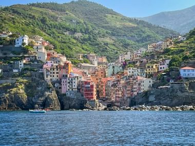Colorful houses of Riomaggiore in Cinque Terre built on rocky cliffs above blue water, with terraced green hillsides in the background.
