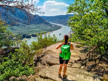 Female hiker with green backpack standing on rocky viewpoint overlooking the Danube River in Wachau Valley with vineyards and mountains.