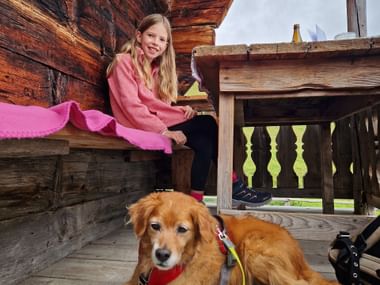 Junges Mädchen in rosa Jacke sitzt auf Holzbank mit Golden Retriever an der Leine bei traditioneller Almhütte in Saalbach, Pinzgau.