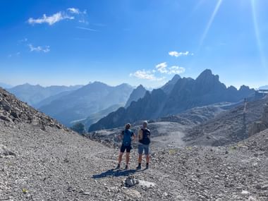 Zwei Wanderer stehen auf felsigem Gelände an der Valluga und blicken auf schroffe Gipfel der Allgäuer und Lechtaler Alpen unter blauem Himmel.