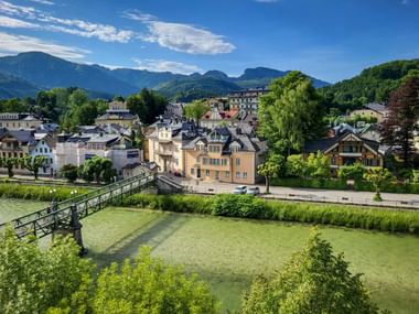 View of Bad Ischl with a green footbridge crossing a river, colorful buildings, and mountain ranges under a blue sky with clouds.