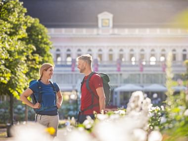 Zwei Wanderer mit Rucksäcken in einem Blumengarten mit dem historischen Kongresshaus in Bad Ischl im Hintergrund.