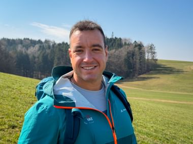 Smiling hiker wearing turquoise jacket with backpack on green hillside near Bozen. Forested hills and blue sky in background.