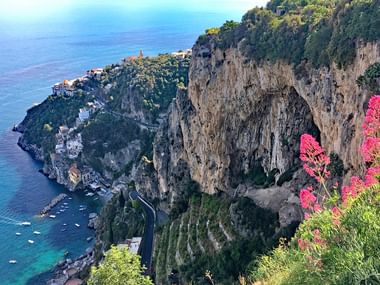 Luftaufnahme von Positano an der Amalfiküste mit bunten Häusern an steilen Klippen, türkisfarbenem Meer, Booten im Hafen und rosa Blüten im Vordergrund.