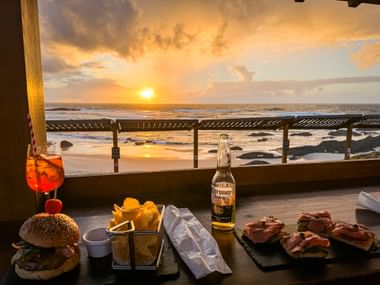 Esstisch mit Burger, Pommes, Sandwiches und Corona-Bier mit Blick auf den Strand Praia de Nossa Senhora bei Sonnenuntergang in Almograve, Alentejo.