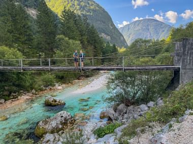 Zwei Wanderer überqueren eine Hängebrücke über dem türkisfarbenen Soča-Fluss in Trenta, Julische Alpen. Berge und Wald im Hintergrund.
