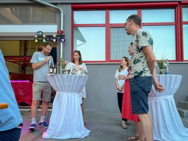 Gruppe von Personen bei Sommerfest im Freien mit weißen Stehtischen. Mann mit Mikrofon spricht zu Gästen vor Gebäude mit roten Fenstern.