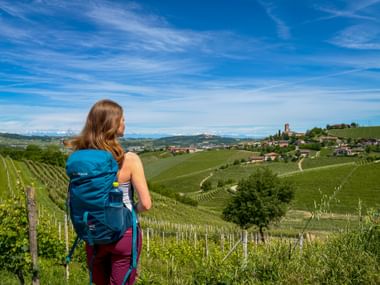 Female hiker with blue backpack viewing rolling vineyard hills in Piedmont, Italy. A hilltop village is visible in the distance.