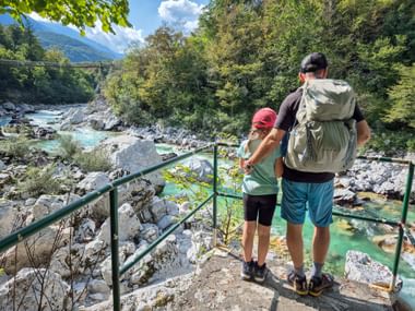 Vater und Kind mit Rucksack stehen auf Aussichtsplattform mit Blick auf türkisfarbenen Soča-Fluss in den Julischen Alpen, umgeben von üppigem Wald.