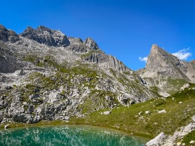 Türkisfarbener Knoppigsee umgeben von grünen Almwiesen und dramatischen Felsgipfeln in den Allgäuer Alpen unter blauem Himmel.