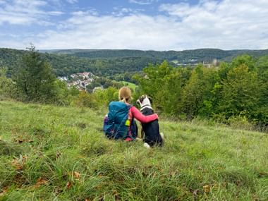 Wanderer mit Rucksack sitzt auf grasigem Hang mit schwarz-weißem Hund und blickt über das Altmühltal mit bewaldeten Hügeln und Dorf.