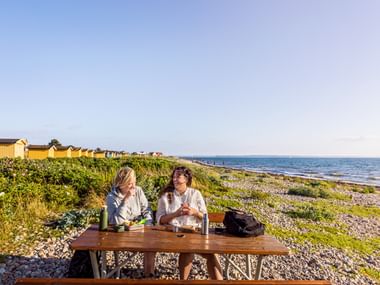 Zwei Frauen picknicken an einem Holztisch am Kieselstrand von Vikens Havsbad. Gelbe Strandhütten und das Meer sind im Hintergrund sichtbar.