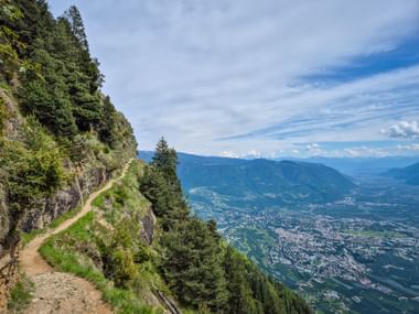 Mountain trail on Meraner Höhenweg winding along a steep slope with panoramic view of Merano town and valley in South Tyrol.