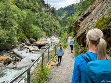 Three hikers walking on a mountain trail along Passer Gorge. A metal railing separates the path from a rocky stream below, with steep cliffs above.