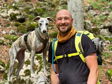 Lächelnder Wanderer mit Rucksack und Windhund auf moosbedeckten Felsen im Wald. Der Hund trägt ein Geschirr, der Mann einen gelben Schal.
