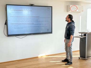 Man in dark polo shirt presenting to a screen showing 'Jahresschwerpunkte 2026' in a bright training room with wooden floor.