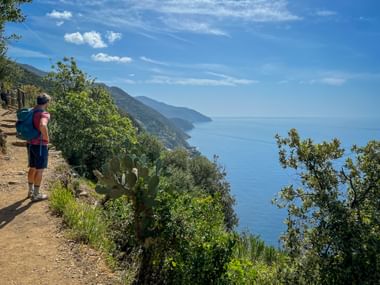 Hiker with backpack on coastal trail in Cinque Terre, overlooking the blue Mediterranean Sea with green hillsides and distant coastline.